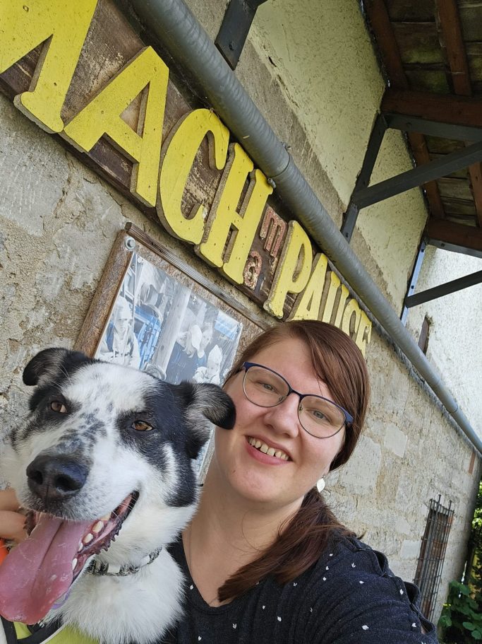 Elene Ruppelt Frau mit Brille lächelt neben einem Hund vor einem Schild mit dem Schriftzug „Mach Pause“.