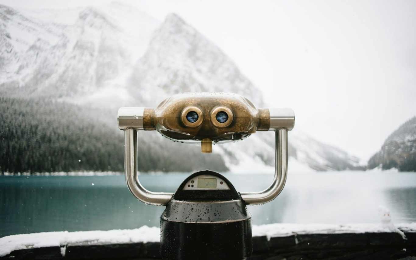 Ein Fernglas steht vor einer schneebedeckten Berglandschaft am Wasser.