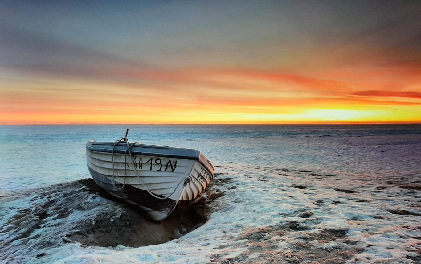 Ein altes Boot liegt im Sand am Ufer, während die Sonne untergeht.